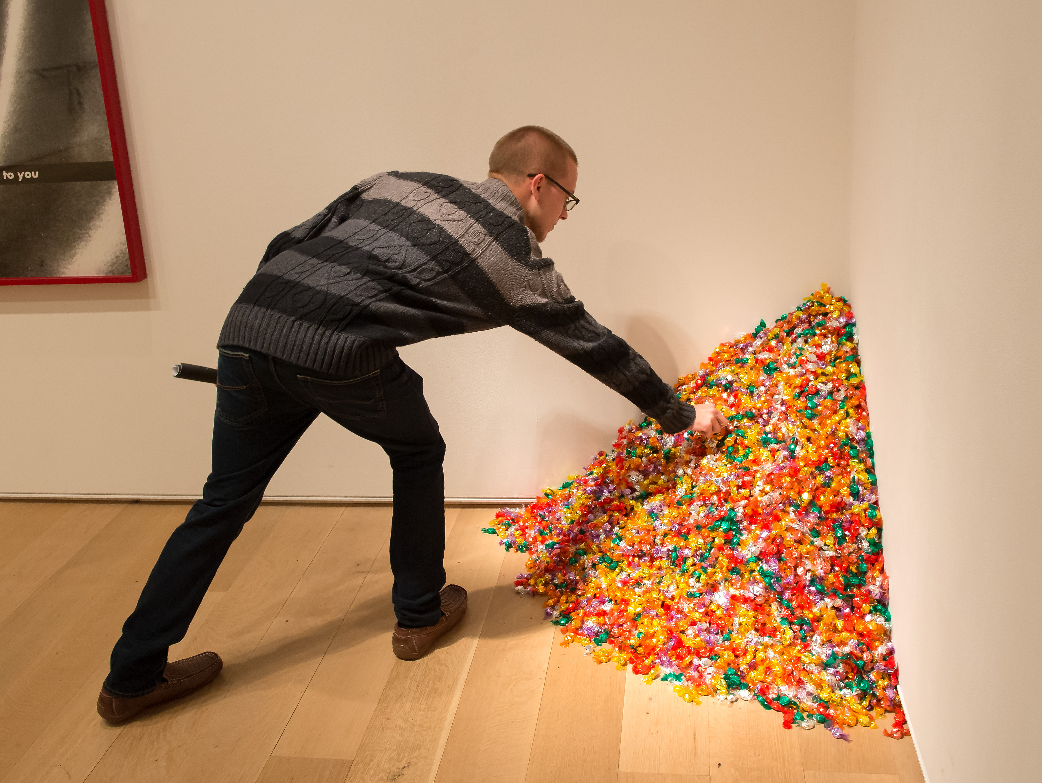 A masculine person with short hair, glasses, and a striped sweater reaching down to grab a piece of candy from a large pile of colorful wrapped candies sitting in the corner of a room. The man has his back turned to the camera